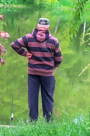 Ein Angler in fortgeschrittenem Alter in Freizeithose, Streifen-Hoodie und mit Schirmmütze am grünen Seeufer.