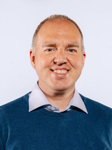 Auf dem Bild ist Arne Bode zusehen. Er hat sehr kurzes Graues Haar und trägt einen blauen Pullover über einem hellblauen Hemd. 