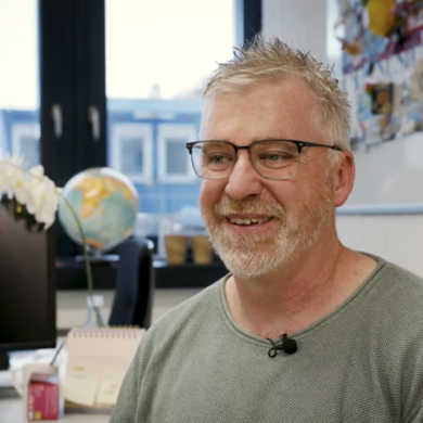 Auf dem Bild ist Robert Rennie zusehen. Er sitzt in einem B&uuml;ro. Der hat kurze graue Haare und tr&auml;gt ein gr&uuml;nes T-Shirt. Im Hintergrund ist ein Globus zusehen. 