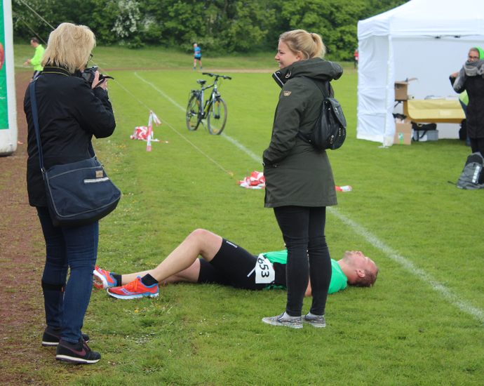 Ein L&auml;ufer liegt ersch&ouml;pft auf den Boden einer Wiese und um ihn herum stehen zwei Frauen.
