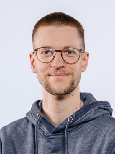 Auf dem Bild ist Patrick Jonasson zusehen. Er hat kurze blonde Haare und einen Bart. Er trägt eine runde Brille und einen dunkelblauen Kaputzenpullover. 