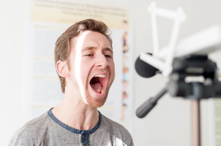 Ein Student spricht mit weit geöffnetem Mund in ein Mikrofon während einer logopädischen Übung.
