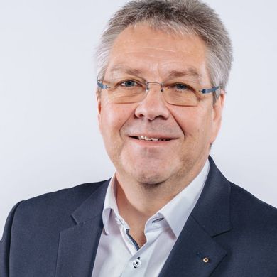 Auf dem Foto ist Helge Ellwart zu sehen. Er hat kurzes graues Haar und trägt ein weißes Hemd unter einer schwarzen Anzugsjacke. 