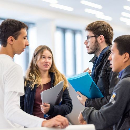 Eine Gruppe Studierender mit Unterlagen in der Hand unterh&auml;lt sich. 