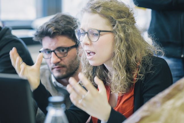 Junge Frau hebt zweifelnd/ emp&ouml;rt die H&auml;nde hoch. Junger Mann sitzt neben ihr. 