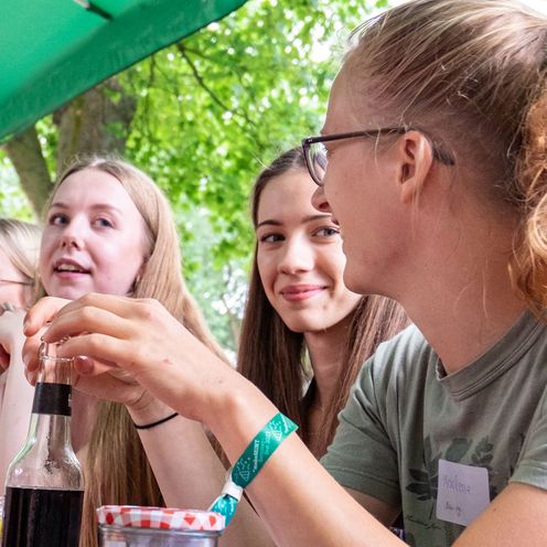 Vier HSB Studierende sitzen drau&szlig;en unter B&auml;umen beim Sommerfest und unterhalten sich. 