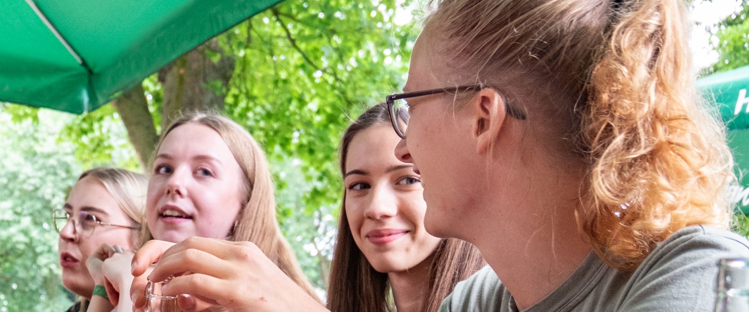 Vier HSB Studierende sitzen drau&szlig;en unter B&auml;umen beim Sommerfest und unterhalten sich. 