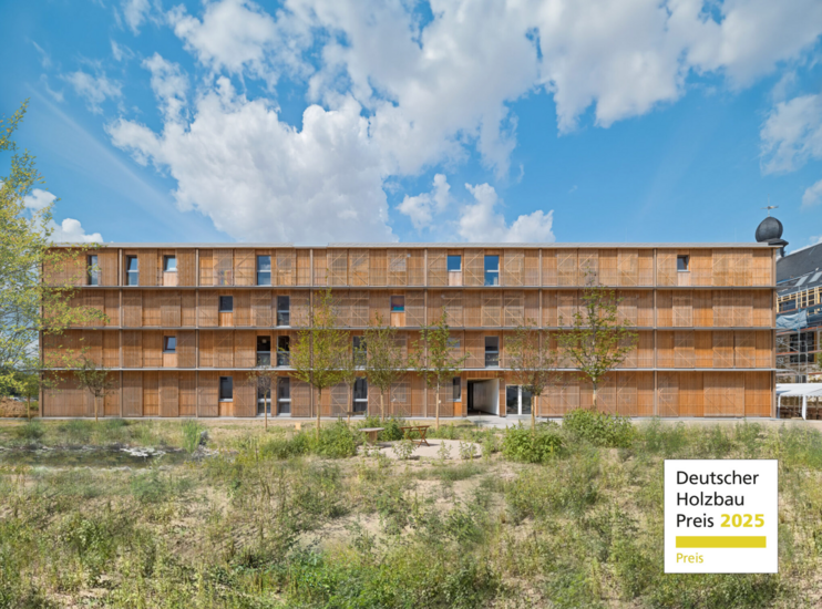 Moderne, vierstöckige Gebäudefassade aus Holz mit vertikalen Lamellen vor den Fenstern, aufgenommen bei blauem Himmel mit weißen Wolken. Vor dem Gebäude befindet sich eine natürliche, begrünte Fläche mit jungen Bäumen und Sitzgelegenheiten. In der rechten unteren Ecke ist ein Logo mit der Aufschrift „Deutscher Holzbau Preis 2025“ zu sehen.