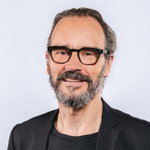 Auf dem Bild ist Jörg Müssig zusehen. Er hat schwarz-grau meliertes Haar und Bart und trägt eine schwarze Brille und ein schwarzes T-Shirt unter einem schwarz Jackett. 