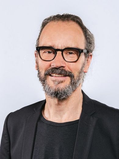 Auf dem Bild ist Jörg Müssig zusehen. Er hat schwarz-grau meliertes Haar und Bart und trägt eine schwarze Brille und ein schwarzes T-Shirt unter einem schwarz Jackett. 