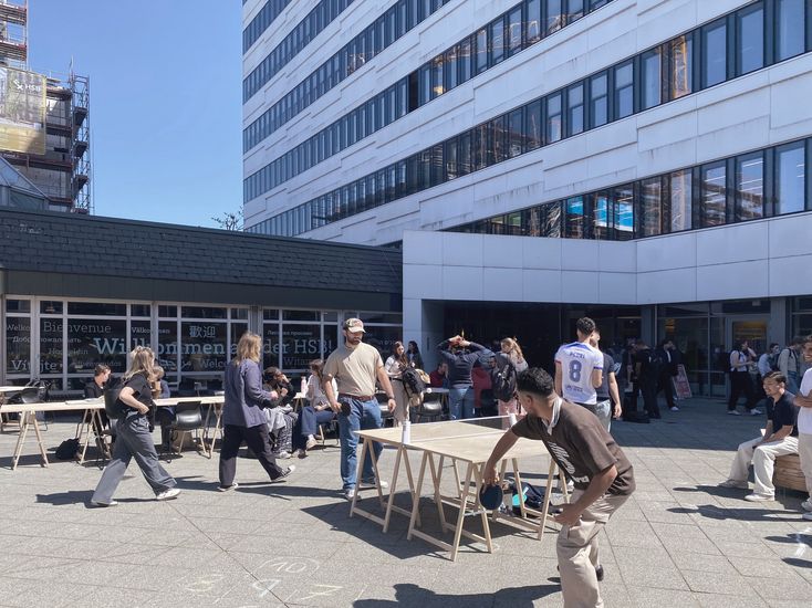 Menschen spielen Tischtennis vor einem Gebäude und sitzen auf Stühlen.