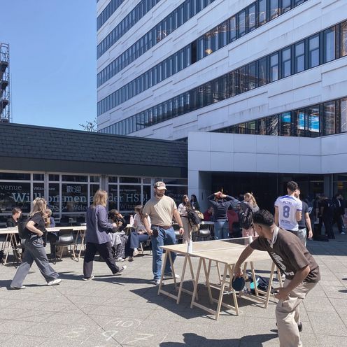 Menschen spielen Tischtennis vor einem Geb&auml;ude und sitzen auf St&uuml;hlen.