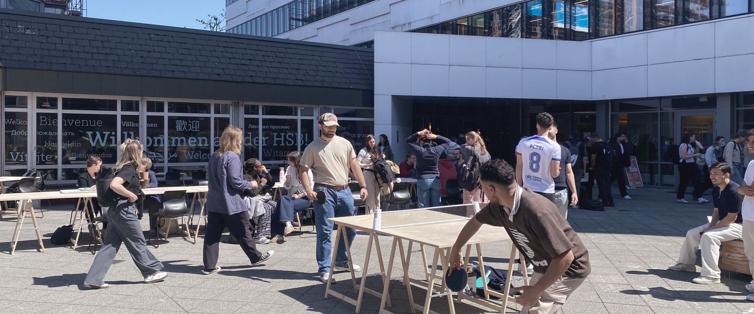 Menschen spielen Tischtennis vor einem Gebäude und sitzen auf Stühlen.