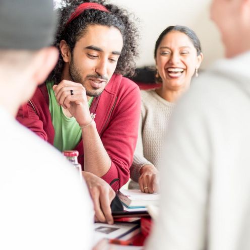 Eine Gruppe von Studierenden sitzt an einem Tisch. Sie lachen und blicken teilweise auf ein Tablet vor ihnen auf dem Tisch.