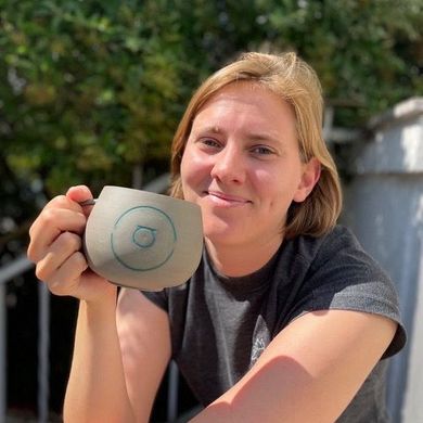 Auf dem Bild ist Annie Berend zusehen. Sie sitzt draußen im Grünen mit einer Tasse in der Hand. Sie trägt ein schwarzes T-Shirt und hat blondes Haar. 