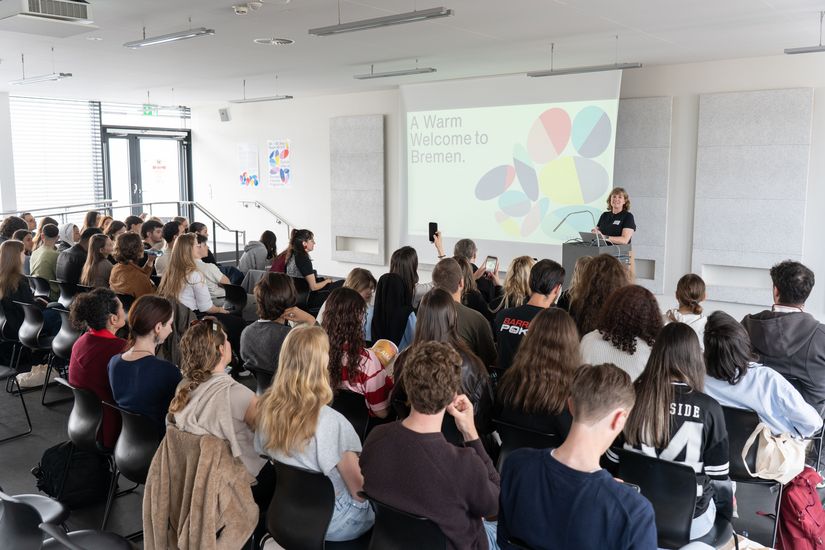 A speaker welcomes a large audience in a brightly lit event hall during a presentation. The slide &lsquo;A Warm Welcome to Bremen&rsquo; is displayed on the screen.