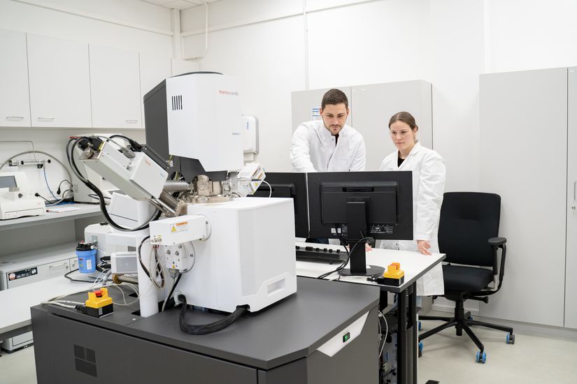 Mann und Frau schauen auf PC. Vor ihnen steht ein gro&szlig;es Mikroskop.