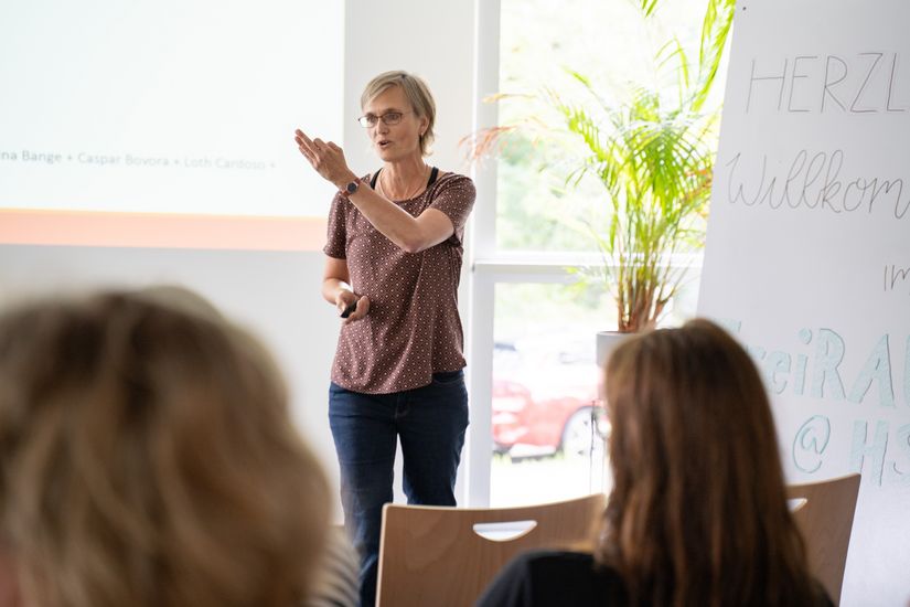 Auf dem Bild steht eine Frau mit kurzen blonden Haaren, die vor einem Publikum gestikuliert und einen Vortrag hält.