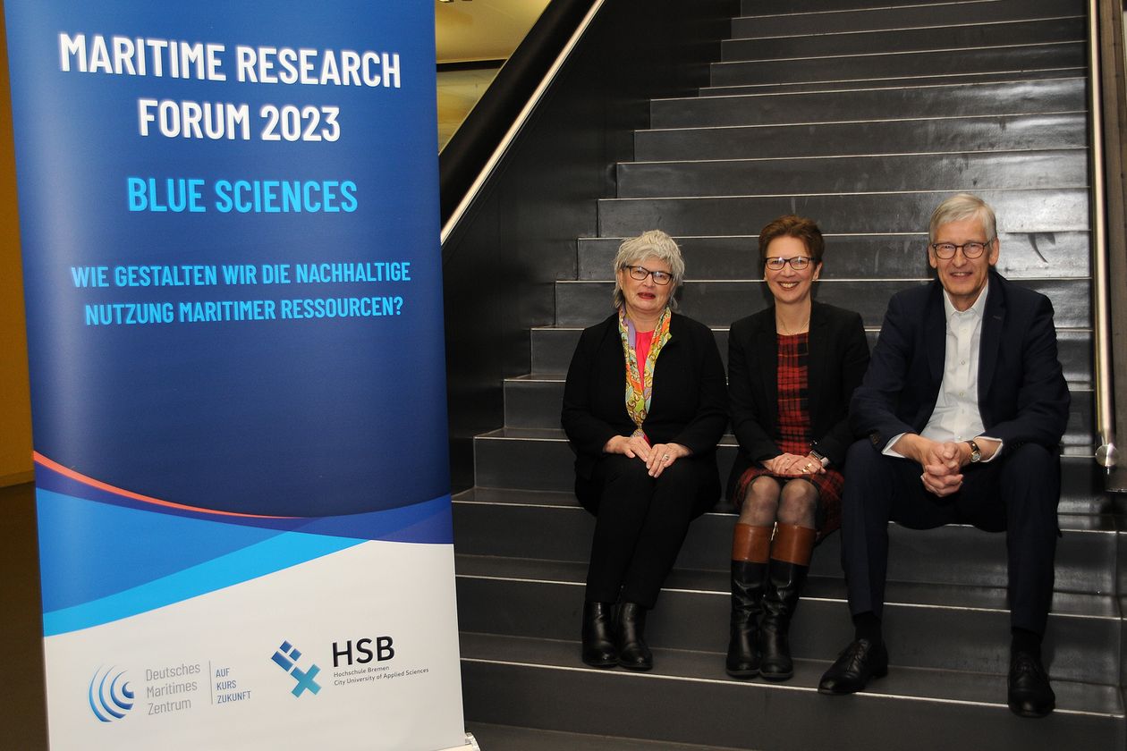 Maritime Research Forum 2023 Gruppenbild mit zwei Frauen und einem Mann. Sie sitzen auf einer Treppe nebeneinander. Neben ihnen steht ein Roll-up.
