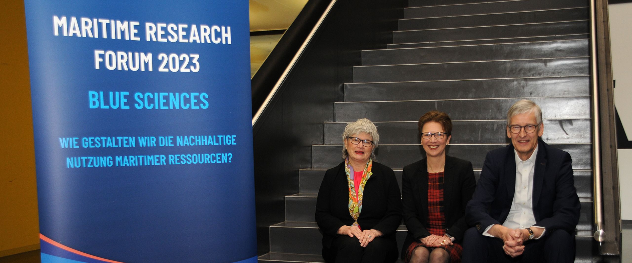 Maritime Research Forum 2023 Gruppenbild mit zwei Frauen und einem Mann. Sie sitzen auf einer Treppe nebeneinander. Neben ihnen steht ein Roll-up.