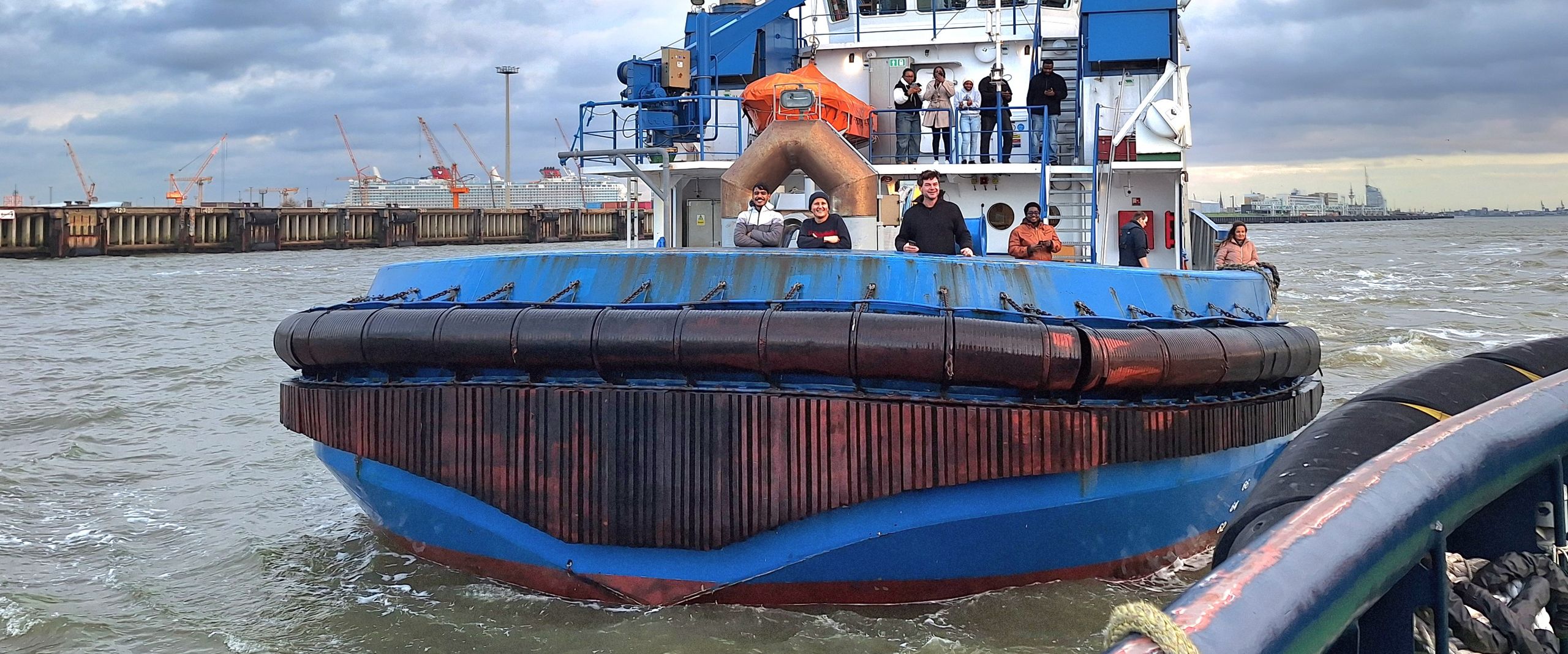 ISSC Exkursion Bremerhaven Zwei Schlepperboote begegnen sich im Hafengelände. Am Bug eines der Schiffe stehen HSB-Studierende und winken in die Kamera.