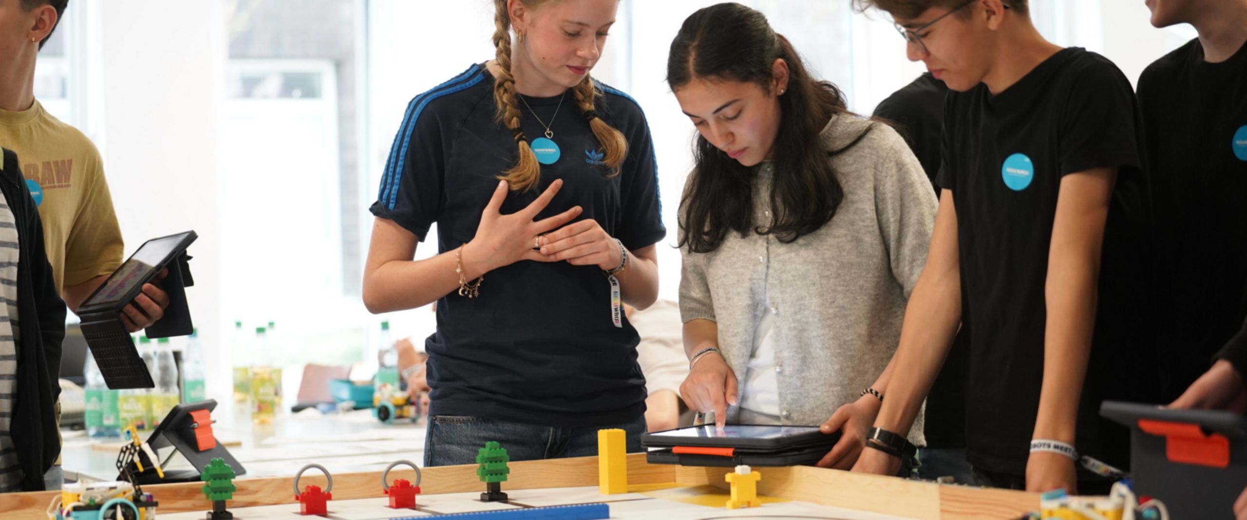 WRO 2026 Mädchen und Jungen stehen an einer buntskizzierten Tischplatte mit Figuren und Roboter.