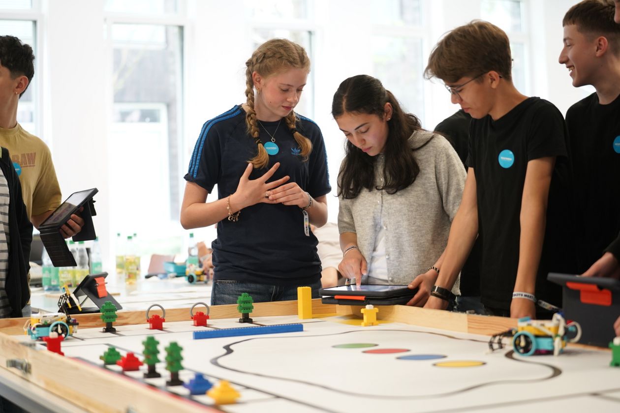 M&auml;dchen und Jungen stehen an einer buntskizzierten Tischplatte mit Figuren und Roboter.