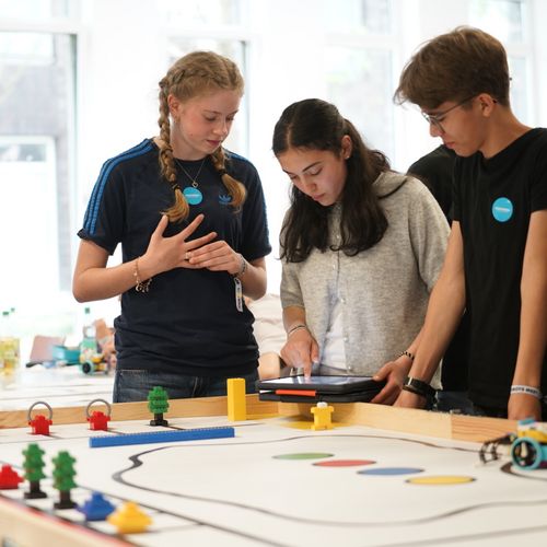 WRO 2026 Mädchen und Jungen stehen an einer buntskizzierten Tischplatte mit Figuren und Roboter.