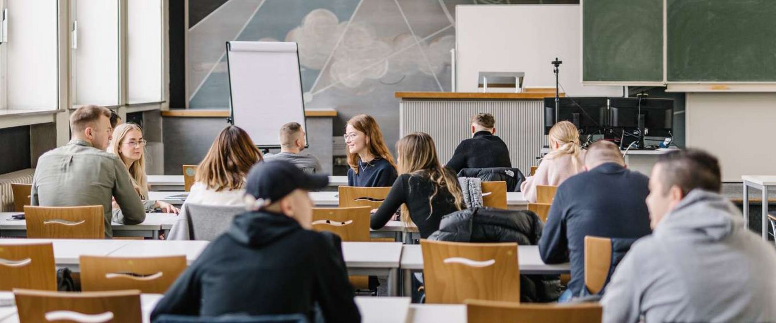 Perspektive aus der hinteren Reihe eines Seminarraums: Studierende sitzen in an mehren Tischreihen verteilt. Einige unterhalten sich miteinander. Vorne im Raum ist eine Leinwand zu sehen, auf die eine Präsentation projiziert ist.