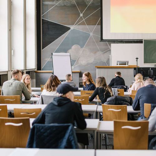 Perspektive aus der hinteren Reihe eines Seminarraums: Studierende sitzen in an mehren Tischreihen verteilt. Einige unterhalten sich miteinander. Vorne im Raum ist eine Leinwand zu sehen, auf die eine Pr&auml;sentation projiziert ist.