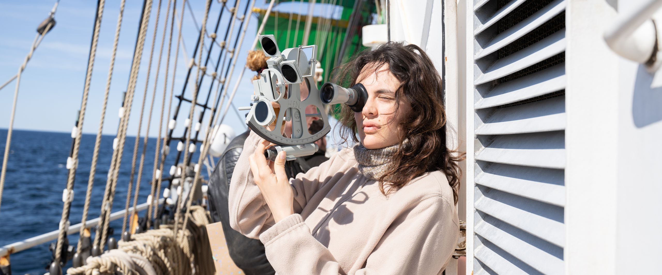 Junge Frau hält Sextanten und befindet sich auf einem Großsegler. Im Hintergrund sieht man Taue.