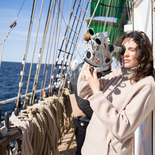 Junge Frau h&auml;lt Sextanten und befindet sich auf einem Gro&szlig;segler. Im Hintergrund sieht man Taue.