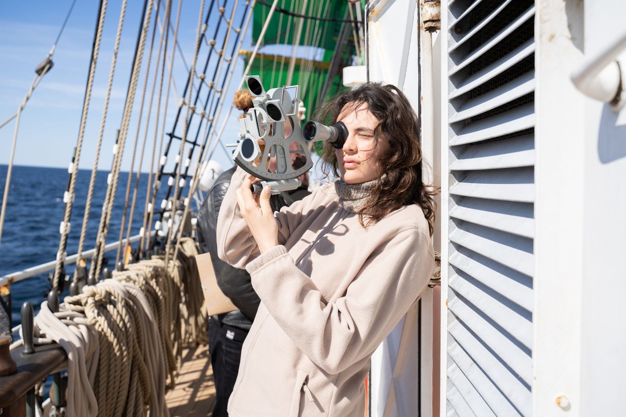Junge Frau hält Sextanten und befindet sich auf einem Großsegler. Im Hintergrund sieht man Taue.