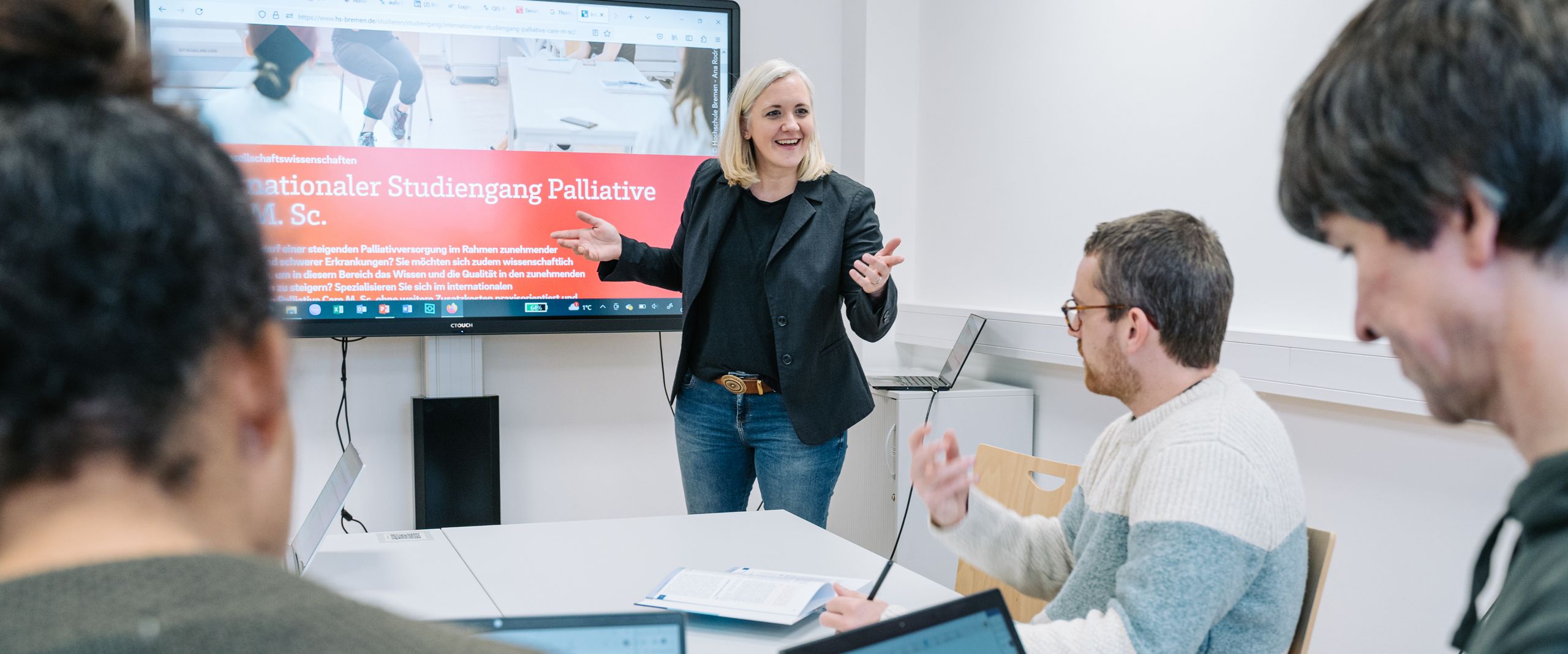 Palliative Care Auf dem Bild ist eine Seminarsituation in kleiner Lerngruppe angemeldet. Die Professorin steht vor einer digitalen Tafel und eine kleine Gruppe Studierende sitzt an Rechnern und hört zu.