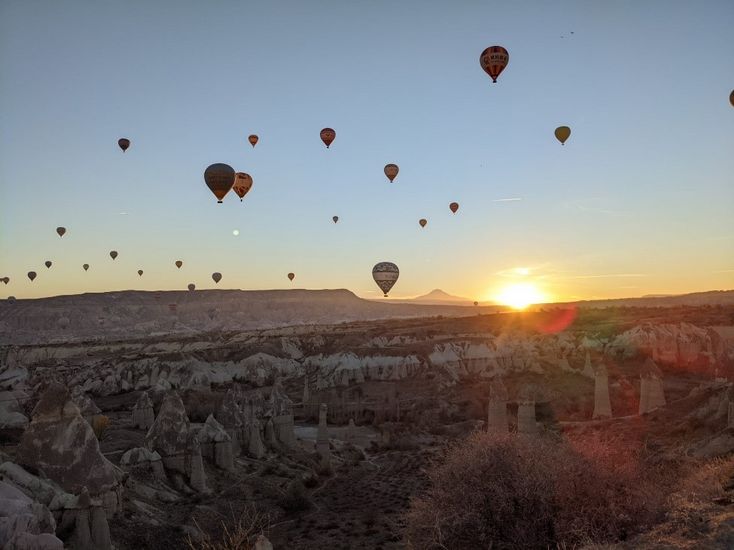 Bild eines Sonnenuntergangs über der hügeligen Landschaft von Kappadokien. Viele Heißluftballone sind am Himmel zu sehen. 