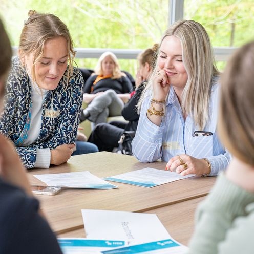 Auf dem Bild sin zwei Studentinnen zusehen, die an einem Tisch mit anderen Studierenden sitzen. Beide sind &uuml;ber ein Aufgabenblatt gebeugt und l&auml;cheln. 