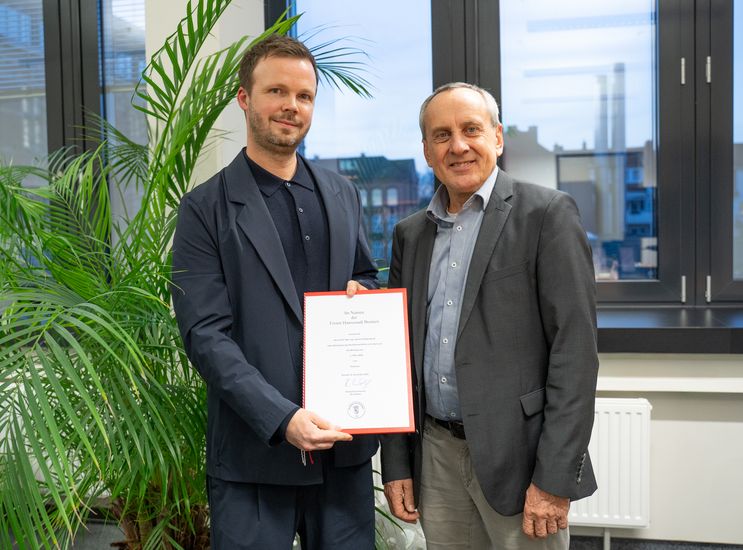 Zwei M&auml;nner schauen in die Kamera. der linke h&auml;lt eine urkunde in den H&auml;nden.