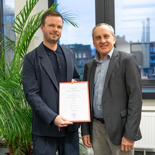 Zwei M&auml;nner schauen in die Kamera. der linke h&auml;lt eine urkunde in den H&auml;nden.