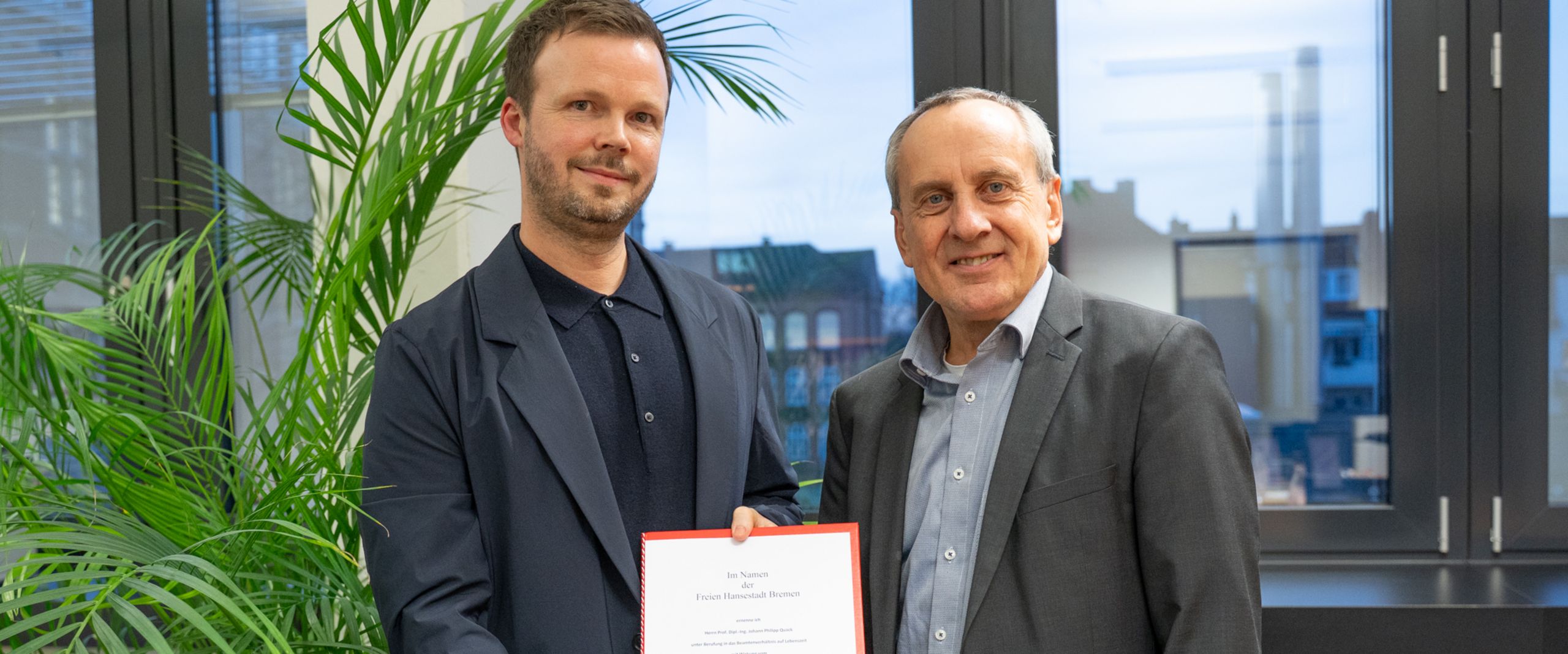 Ernennung Philipp Quack Zwei Männer schauen in die Kamera. der linke hält eine urkunde in den Händen.