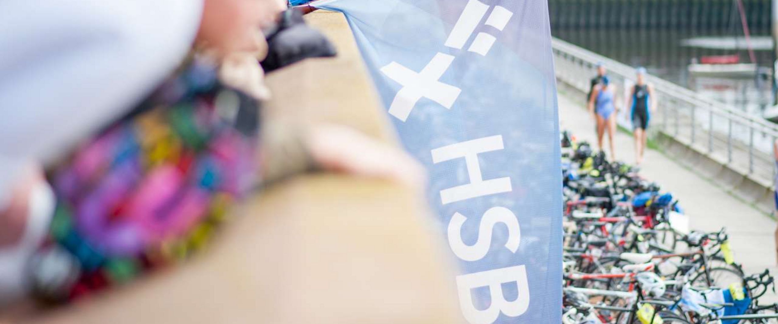 Fahne HSB Gewoba Triathlon Eine blaue Fahne mit dem HSB Logo wird von einem Mann gehalten. Im Hintergrund ist der Marina Hafen in Bremen-Überseestadt zu sehen.