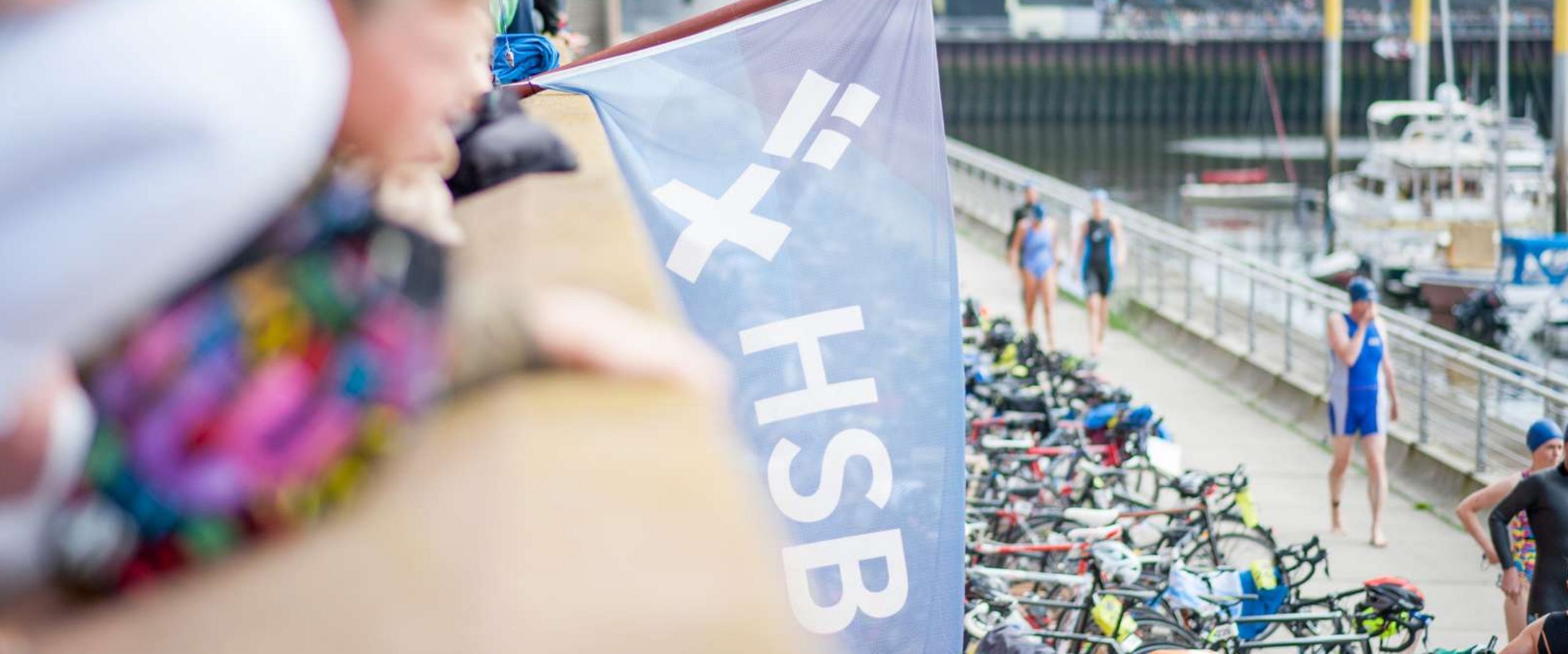 Fahne HSB Gewoba Triathlon Eine blaue Fahne mit dem HSB Logo wird von einem Mann gehalten. Im Hintergrund ist der Marina Hafen in Bremen-Überseestadt zu sehen.