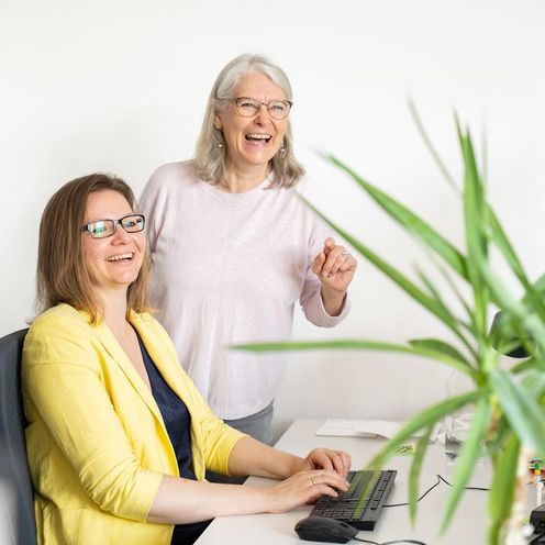Auf dem Foto sind zwei Frauen zu sehen. Eine sitzt an einem Schreibtisch vor einem Computer, die andere steht hinter dem Stuhl. Rechts im Bild ist eine gr&uuml;ne Palme. Beide Frauen lachen und schauen knapp an der Kamera vorbei. 