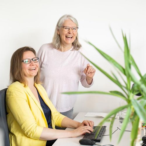 Auf dem Foto sind zwei Frauen zu sehen. Eine sitzt an einem Schreibtisch vor einem Computer, die andere steht hinter dem Stuhl. Rechts im Bild ist eine gr&uuml;ne Palme. Beide Frauen lachen und schauen knapp an der Kamera vorbei. 