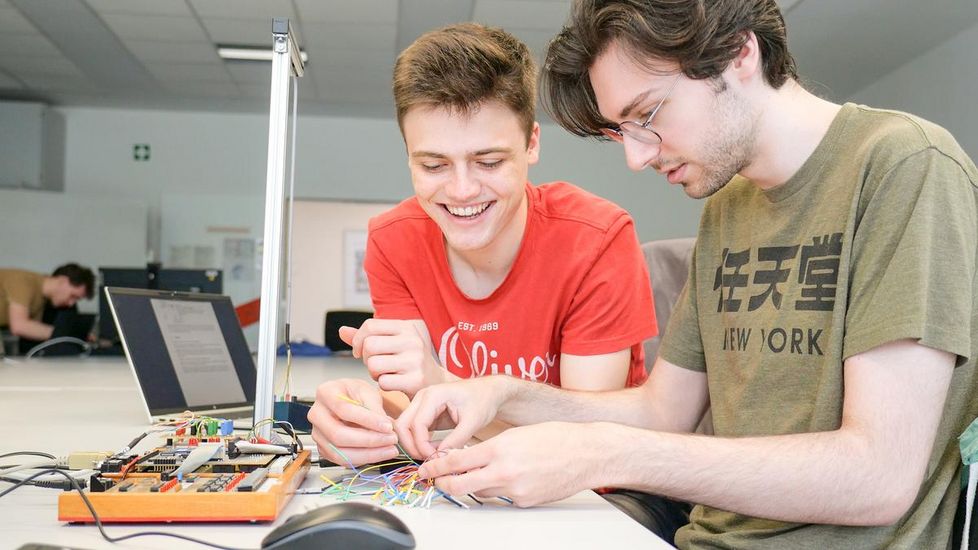 Auf dem Bild sind zwei Studenten zusehen. Sie sitzen an einem weißen Tisch vor vielen verschiedenen bunten Kabeln und einem Laptop. Beide lachen. 
