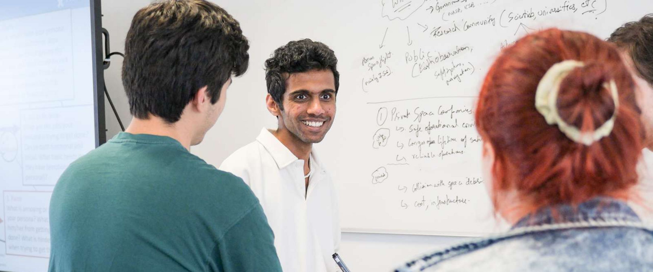 Auf dem Bild ist eine Gruppe Studierender zusehen. Sie stehen vorne an einer Tafel. Ein Student blickt knapp an der Kamera vorbei und lacht. Er hat einen Stift in der Hand um etwas an die Tafel schreiben zu können. Die anderen Studierenden sind nur unscharf zu erkennen. 