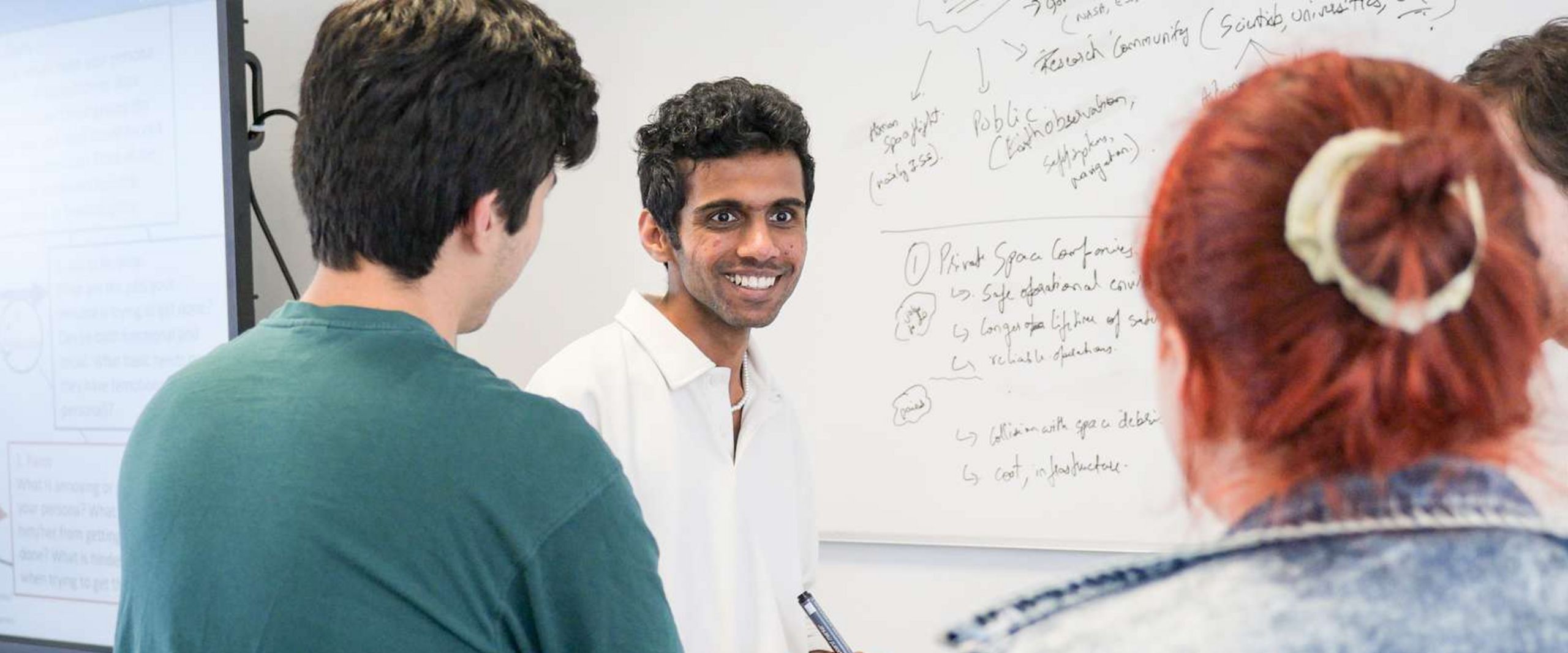 Auf dem Bild ist eine Gruppe Studierender zusehen. Sie stehen vorne an einer Tafel. Ein Student blickt knapp an der Kamera vorbei und lacht. Er hat einen Stift in der Hand um etwas an die Tafel schreiben zu können. Die anderen Studierenden sind nur unscharf zu erkennen. 
