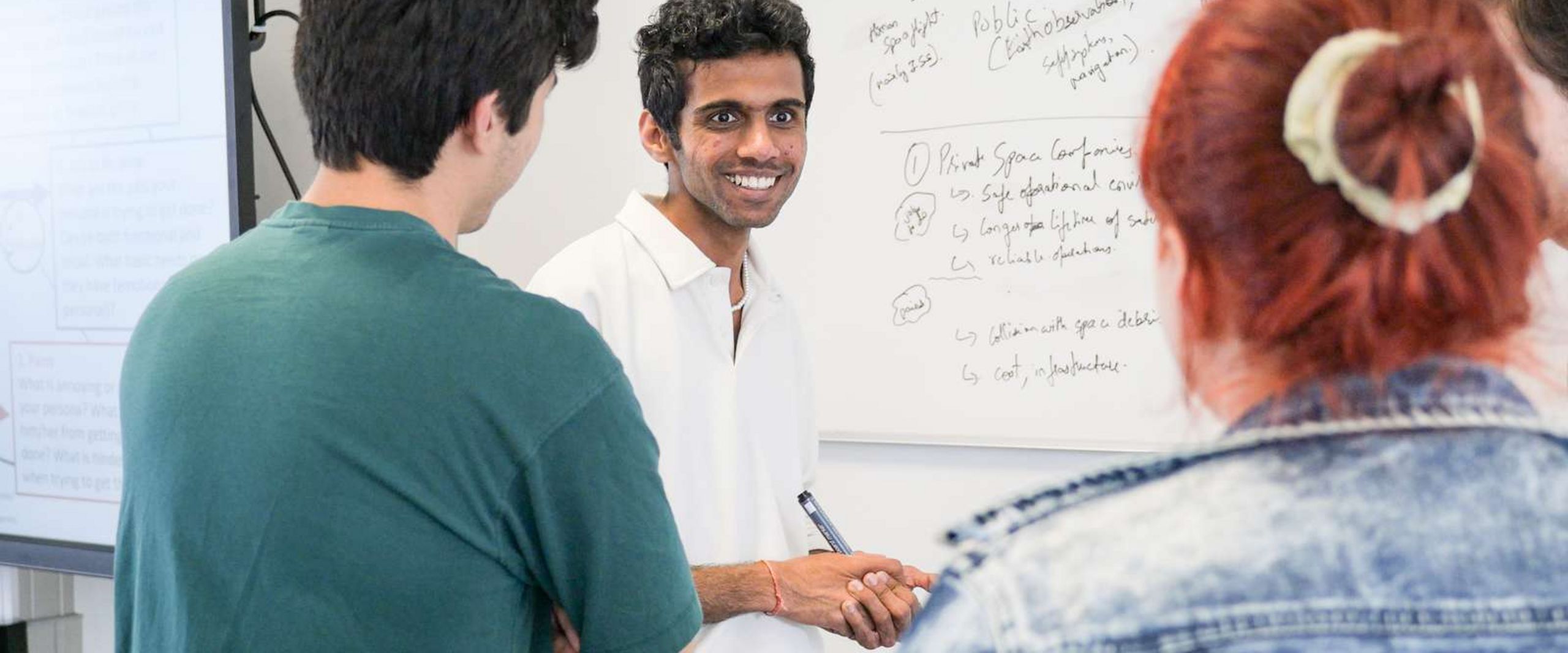 Gruppenarbeit Auf dem Bild ist eine Gruppe Studierender zusehen. Sie stehen vorne an einer Tafel. Ein Student blickt knapp an der Kamera vorbei und lacht. Er hat einen Stift in der Hand um etwas an die Tafel schreiben zu können. Die anderen Studierenden sind nur unscharf zu erkennen.