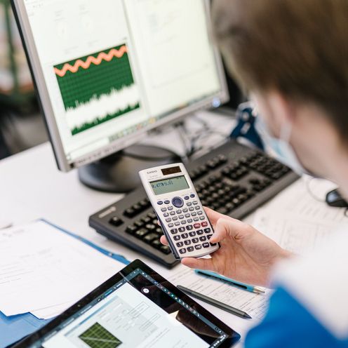Eine Person arbeitet mit einem Taschenrechner, einem Computer und einem Tablet.