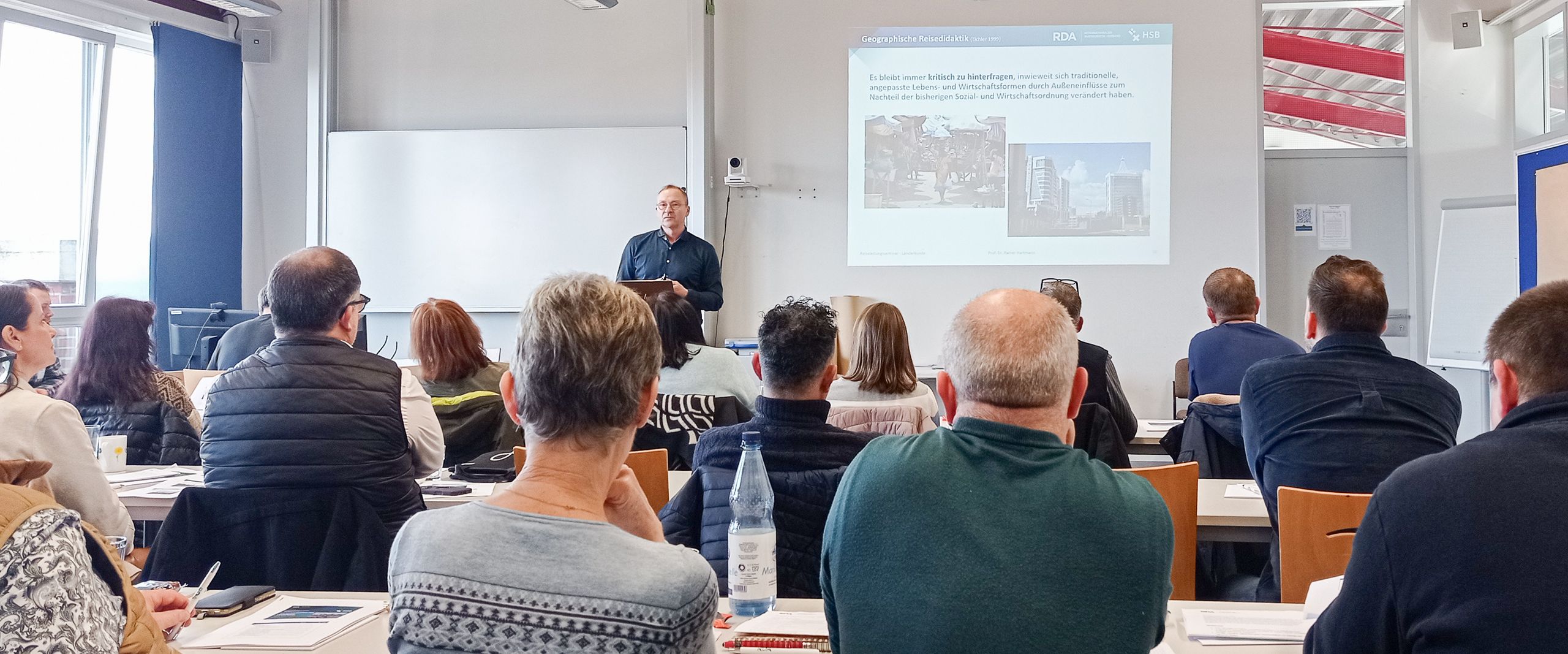Reiseleiterseminar 2025 Auf dem Bild sitzen viele Menschen in einem Raum, den Blick nach vorne gerichtet. Vorne steht ein Mann, der einen Vortrag hält.