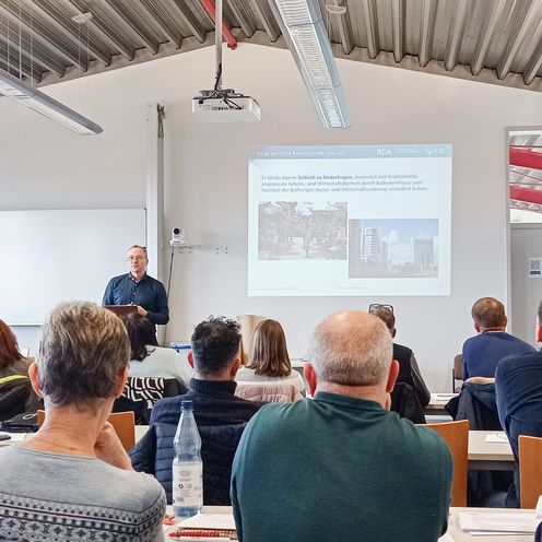 Auf dem Bild sitzen viele Menschen in einem Raum, den Blick nach vorne gerichtet. Vorne steht ein Mann, der einen Vortrag h&auml;lt. 
